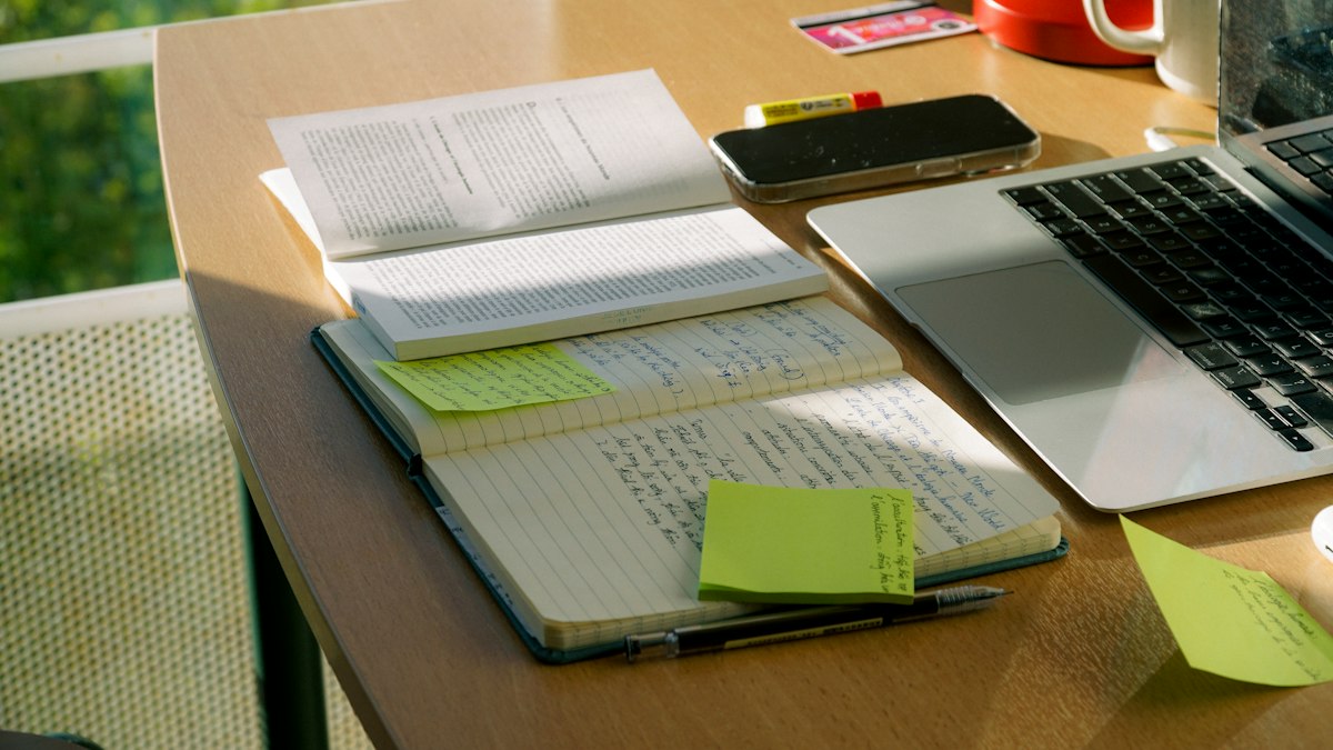 Libro, quaderno di appunti e computer su una scrivania accanto a una finestra.